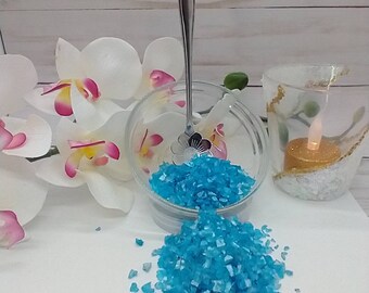 May include: A glass bowl filled with turquoise blue decorative glass crystals, with a silver spoon resting on top. The bowl is sitting on a white surface with white orchids in the background.