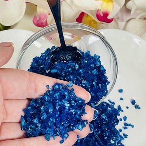 May include: A glass bowl filled with blue iridescent glass pieces. A hand is holding a small amount of the glass pieces.