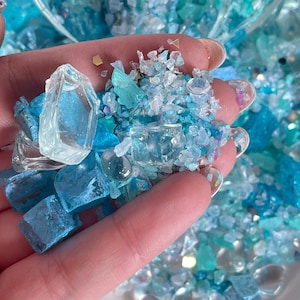 May include: A close-up of a hand holding a variety of blue and clear glass pieces. The pieces are of different shapes and sizes, and some have a sparkly finish.