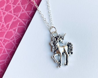 May include: Silver-toned unicorn pendant necklace. The unicorn charm is detailed with a mane, tail, and horn. The necklace is displayed on a white and pink textured background. A delicate chain completes the necklace.