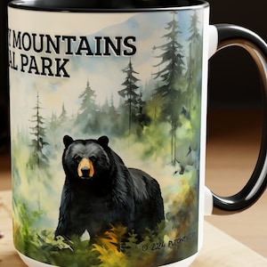 May include: White ceramic mug with a black handle and rim. The mug features a watercolor illustration of a black bear in a misty forest with the text "Smoky Mountains National Park".
