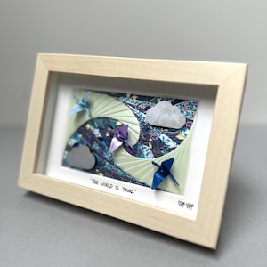 May include: A framed 3D paper art piece featuring three origami cranes in shades of light blue, purple, and dark blue flying over layered patterned paper waves. Two glittery clouds are included. The text reads 'THE WORLD IS YOURS' with a signature.