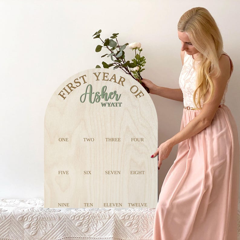 May include: First Year Milestone Arch Sign.  A light beige wooden arch-shaped sign with the text 'First Year Of Asher Wyatt' in brown and green. Below are twelve numbered sections for monthly photos.  The sign is a great keepsake for a baby's first year.