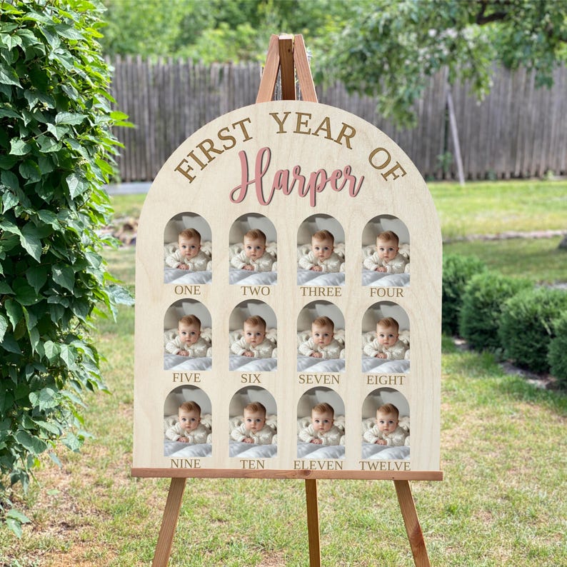 May include: Wooden first-year photo display features twelve arched openings for photos, each labeled with a month.  The top arch displays 'First Year of Harper' in elegant gold and rose-gold lettering.  The light-beige wood provides a neutral backdrop for the baby's pictures.  This charming keepsake is perfect for capturing a child's first year milestones.