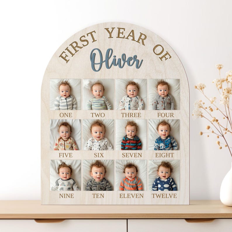 May include: Wooden arched first-year photo display features twelve monthly baby photos in a grid layout.  Each photo is labeled with the month number (one through twelve).  The top of the arch is inscribed with 'FIRST YEAR OF Oliver' in a serif font.  The text is in shades of blue and gold on a light wood background.