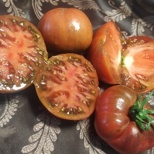 May include: Five heirloom tomatoes, some sliced in half, showing their red flesh and dark brown skin. The tomatoes are on a dark brown fabric with a floral pattern.