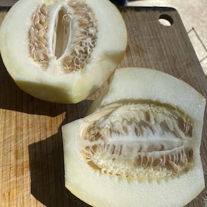 Puede incluir: Dos mitades de melón cantalupo sobre una tabla de cortar de madera. La pulpa de color amarillo pálido del melón es visible, junto con las semillas y la cáscara marrones. Las superficies cortadas muestran la estructura interna del melón.