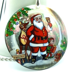 May include: A round, iridescent, painted glass ornament with a depiction of Santa Claus, a reindeer, an elf, and a sign that reads "North Pole".