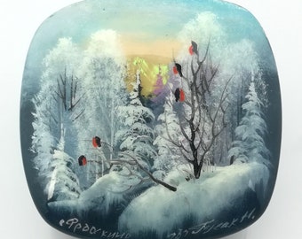 May include: A round, blue, glass trinket box with a winter scene painted on the top. The scene depicts a snowy forest with a few red birds perched on a tree branch. The box is signed "Фроскино" and "937 Freak H."