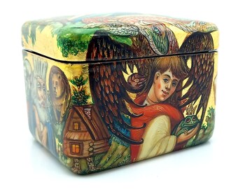 May include: A wooden box with a gold background and a painted scene of a winged figure holding a frog. The scene also includes a small house and other figures.