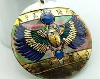 May include: A circular pendant with an Egyptian scarab beetle design. The pendant has a mother-of-pearl base with a painted scarab beetle in gold, blue, and green. It hangs from a black cord with a silver-coloured clasp.