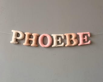 May include: A banner spelling out the name "PHOEBE" in soft, plush letters. The letters are in varying shades of cream, peach, and pink, strung together on a thin, nearly invisible string against a gray background. The letters are puffy and three-dimensional.