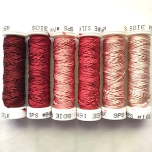 May include: Six spools of embroidery floss in shades of red, pink, and beige. The spools are labelled with the brand name "SOIE" and the colour number. The spools are arranged in a gradient from darkest to lightest.