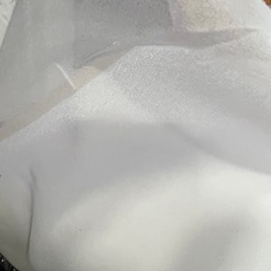 Op de afbeelding: Een close-up van een witte, getextureerde stof. De stof lijkt zacht en mogelijk gelaagd, met subtiele variaties in licht en schaduw. De algemene indruk is van een schoon, ongerept materiaal.