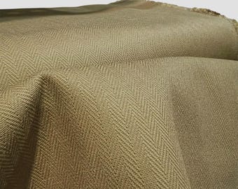 May include: Close-up of a tan-colored fabric with a herringbone weave pattern. The fabric is folded, creating shadows and highlighting the texture. The edges of the fabric are visible, showing a raw, unfinished edge. The overall appearance suggests a textile product.