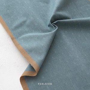 May include: Close-up of a folded, teal fabric with a subtle cross pattern. The fabric has a tan border and the word "FABLEISM" is printed in white. The fabric appears soft and textured, suitable for apparel or home decor.