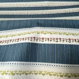 May include: Close-up of a fabric with a striped pattern in shades of blue, white, and green. The design includes rows of vertical stripes and horizontal bands with floral and dotted details. The fabric has a soft, textured appearance.