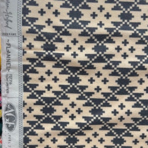May include: A close-up of a cream-colored fabric with a black and white geometric pattern. The pattern features a repeating design of squares and crosses. The fabric is soft and has a slightly textured surface.