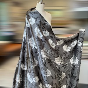 May include: A bolt of fabric draped over a mannequin. The fabric is a dark gray with a floral pattern of white and gray flowers and leaves. The fabric has a slight sheen.