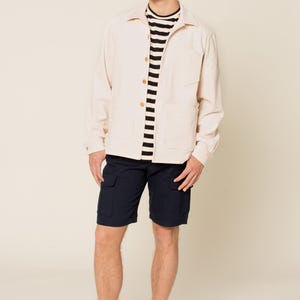 May include: A man wearing a white corduroy jacket over a black and white striped shirt, dark blue cargo shorts, and white sneakers.