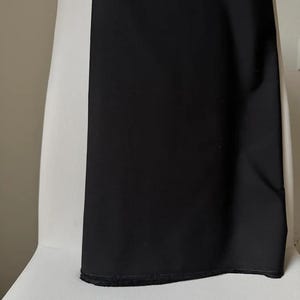 May include: A black fabric, possibly a garment, is draped over a white surface. The fabric is smooth and opaque, with a visible hem at the bottom. The contrast between the black fabric and the white surface creates a striking visual effect.