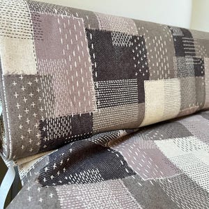 May include: A rolled-up fabric with a patchwork design in shades of gray, brown, and beige. The fabric features various stitched patterns, including crosses and dashes, creating a textured appearance. The design is reminiscent of a quilt or a decorative textile.
