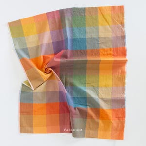 May include: A colorful fabric with a large plaid pattern. The fabric features a variety of colors, including orange, yellow, blue, and purple. The fabric is folded and has the word "FABLEISM" printed on it.