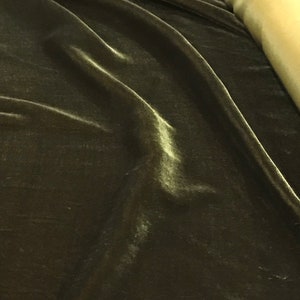 May include: Close-up of dark olive green velvet fabric with subtle highlights and folds. A roll of light gold fabric is visible in the background. The velvet has a soft, luxurious appearance.