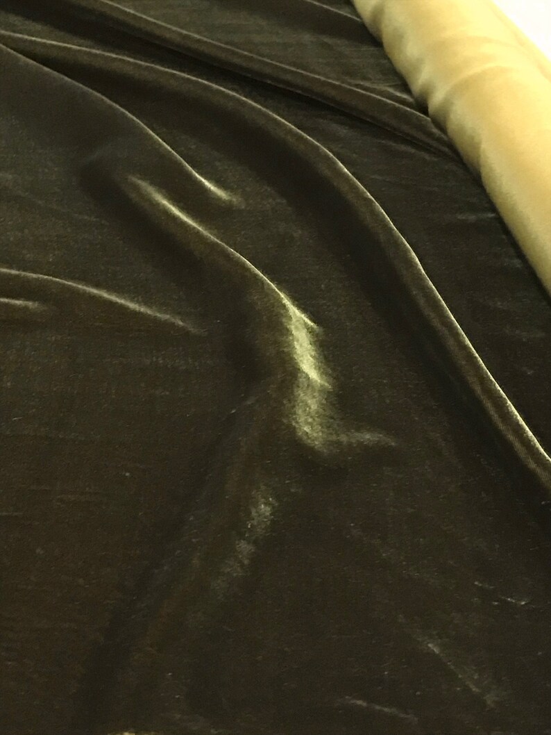 May include: Close-up of dark olive green velvet fabric with subtle highlights and folds. A roll of light gold fabric is visible in the background. The velvet has a soft, luxurious appearance.