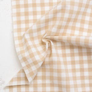 May include: Tan and white gingham fabric with a soft, wrinkled texture. The fabric is folded and draped, showcasing the classic checkered pattern. The image is well-lit, highlighting the fabric's details.