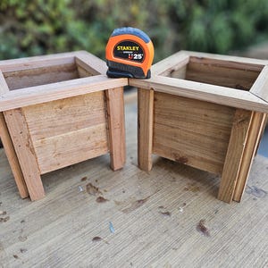 Könnte beinhalten: Zwei unfertige Holzpflanzkästen mit rechteckigem Design. Ein schwarz-oranges Maßband, markiert mit "1 25'", liegt zwischen ihnen. Die Pflanzkästen sind aus hellbraunem Holz und stehen auf einer Holzoberfläche.