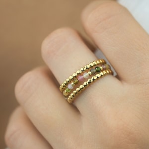 May include: Gold ring with three bands of small gold beads and colourful gemstones. The gemstones are pink, green, and white.