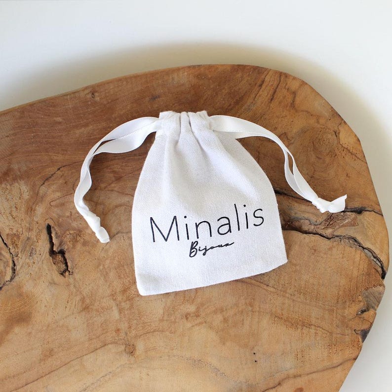 May include: A small, white drawstring bag with the text "Minalis Bijoux" printed in black. The bag is made of a soft fabric and has white ribbon ties. It rests on a textured, wooden surface.