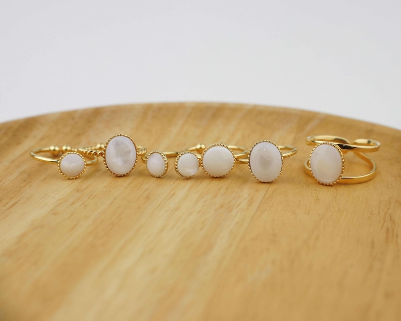 May include: Set of five gold rings with white oval stones. The rings are arranged on a wooden surface.