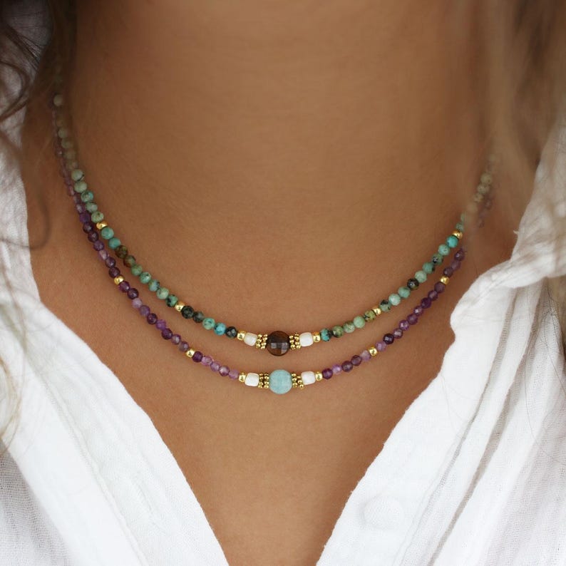 May include: Two layered beaded necklaces. The top necklace features turquoise and gold beads. The bottom necklace has purple and gold beads. Both necklaces have a central bead. The necklaces are worn on a person wearing a white shirt.