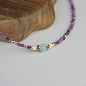 May include: Delicate necklace featuring small, faceted purple beads interspersed with gold-colored accents. A central light blue gemstone is flanked by white beads and gold details, creating a refined and elegant jewelry piece.