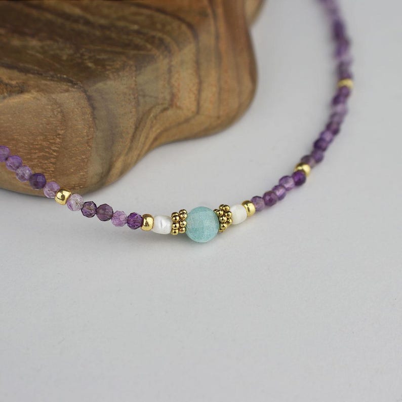 May include: Delicate necklace featuring small, faceted purple beads interspersed with gold-colored accents. A central light blue gemstone is flanked by white beads and gold details, creating a refined and elegant jewelry piece.
