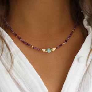 Peut inclure: Délicat collier de perles avec des perles d'améthyste et dorées. Le collier comporte une pierre d'aigue-marine et des perles blanches. Le collier est porté sur une chemise blanche.