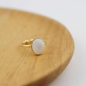 May include: A gold ring with a white oval-shaped stone set in a gold bezel. The ring is on a wooden surface.