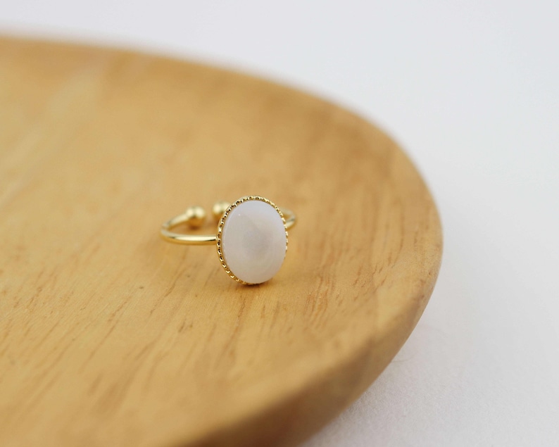 May include: A gold ring with a white oval-shaped stone set in a gold bezel. The ring is on a wooden surface.