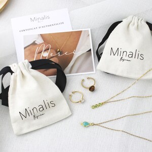 May include: Two white drawstring pouches with black ribbon ties and the text "Minalis Bijoux" printed on them. The pouches are filled with gold necklaces and rings with green and brown stones.