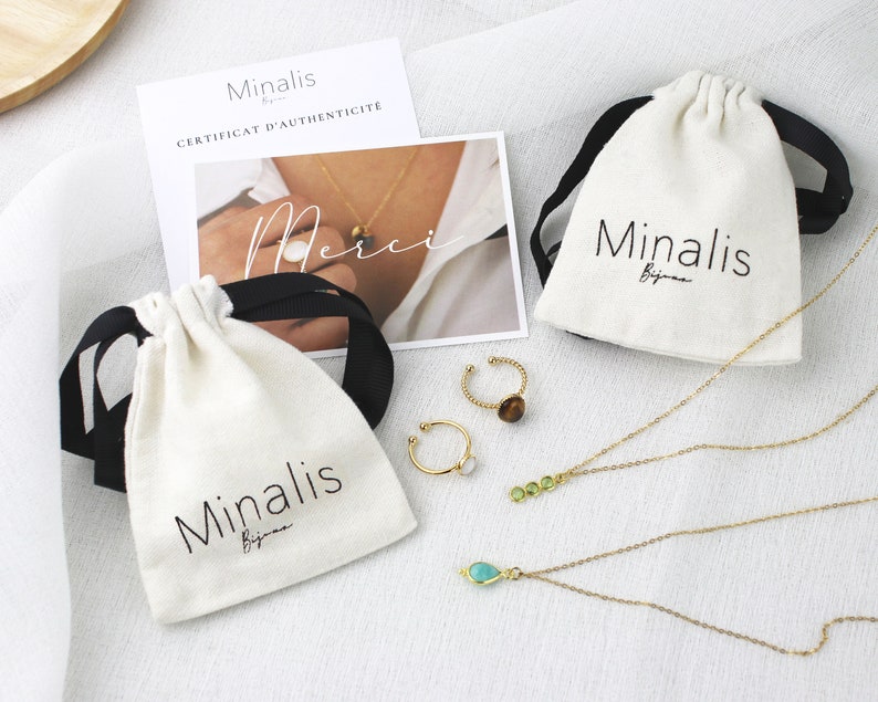 May include: Two white drawstring pouches with black ribbon ties and the text "Minalis Bijoux" printed on them. The pouches are filled with gold necklaces and rings with green and brown stones.