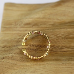 May include: A gold-toned ring with a beaded design featuring small, colourful gemstones. The ring is on a wooden surface.
