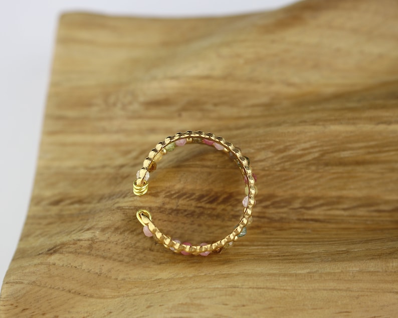 May include: A gold-toned ring with a beaded design featuring small, colourful gemstones. The ring is on a wooden surface.
