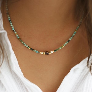 May include: A delicate necklace featuring small turquoise and gold beads. The necklace has a central focal point with a brown bead and two white beads. The necklace is worn around the neck.