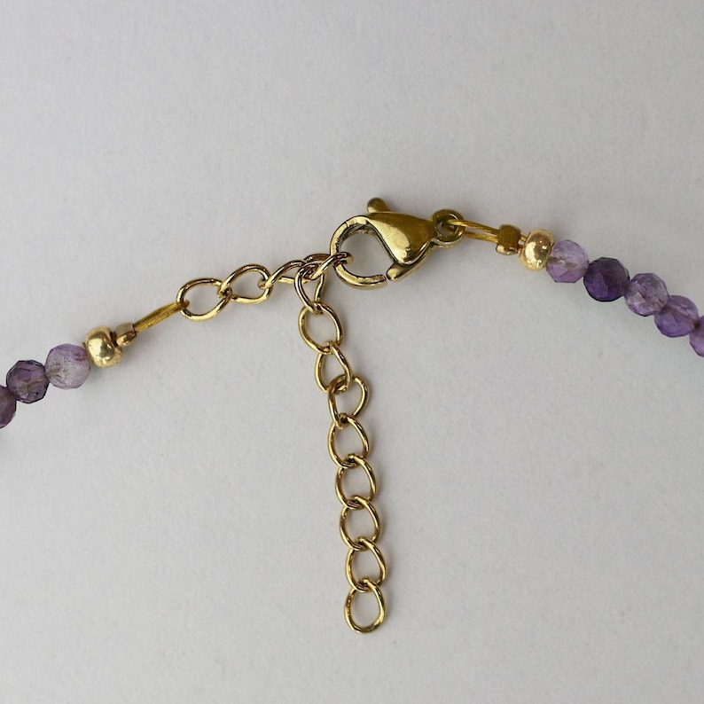 May include: Close-up of a necklace clasp and chain. The necklace features small, round, purple beads. The clasp and chain are gold-colored. The necklace is on a white background.