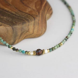 May include: Delicate beaded necklace featuring turquoise and gold-colored beads. The necklace has a central brown bead, with white and gold accents. The necklace is displayed on a wooden surface.