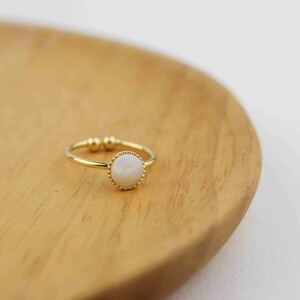 May include: A gold ring with a white pearl-like stone set in a gold bezel. The ring is on a wooden surface.