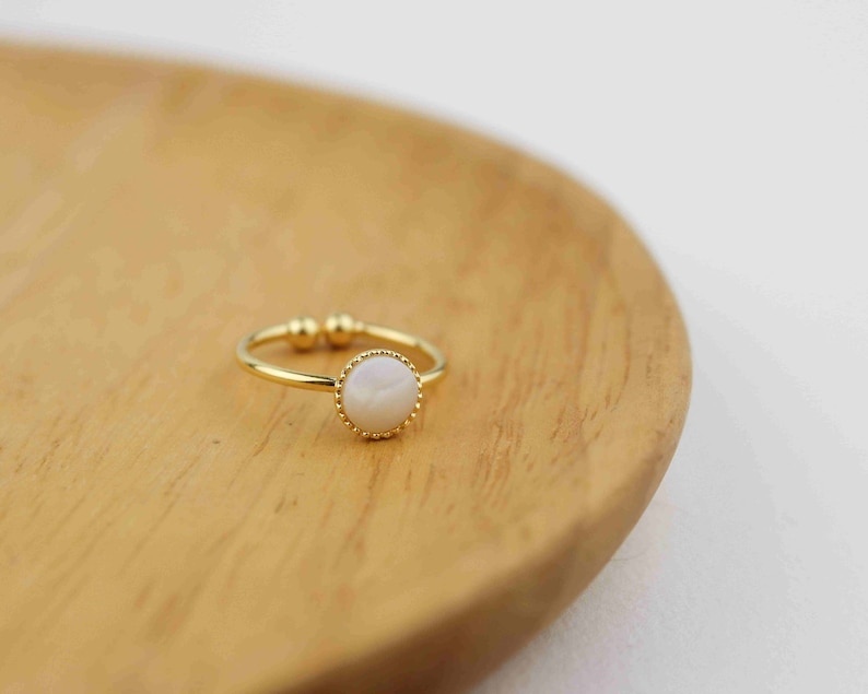 May include: A gold ring with a white pearl-like stone set in a gold bezel. The ring is on a wooden surface.