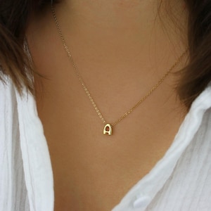 Pode incluir: Um delicado colar de ouro com um pingente com a letra "A". O pingente é pequeno e está centrado em uma corrente fina. O colar é usado sobre uma camisa branca.